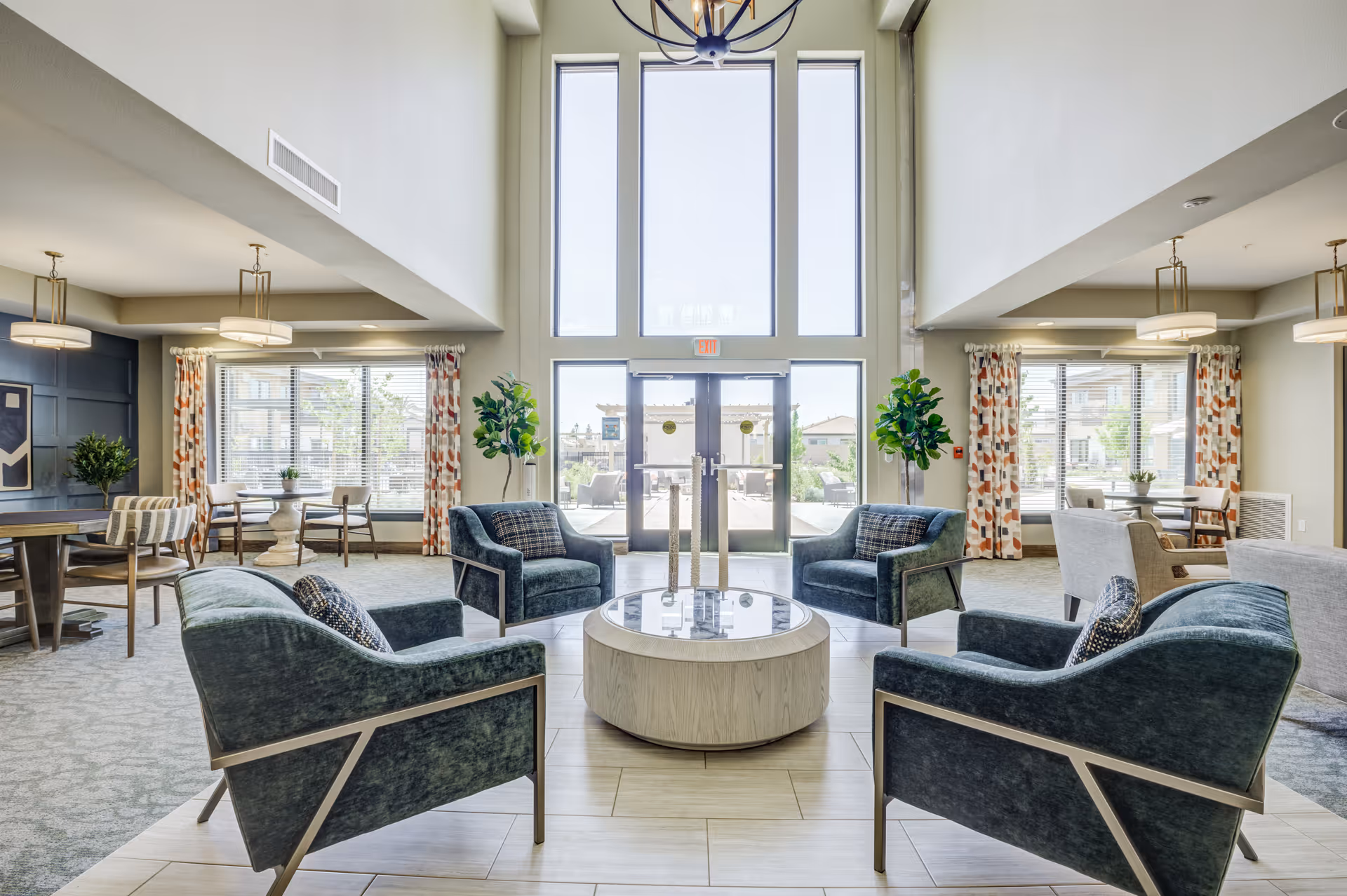 A bright and spacious senior living facility common area with four modern blue armchairs arranged around a round wooden coffee table. Large floor-to-ceiling windows and glass doors let in natural light, with patterned curtains on the side windows. There are additional seating areas with tables and chairs visible in the background, along with decorative plants and ceiling lights.