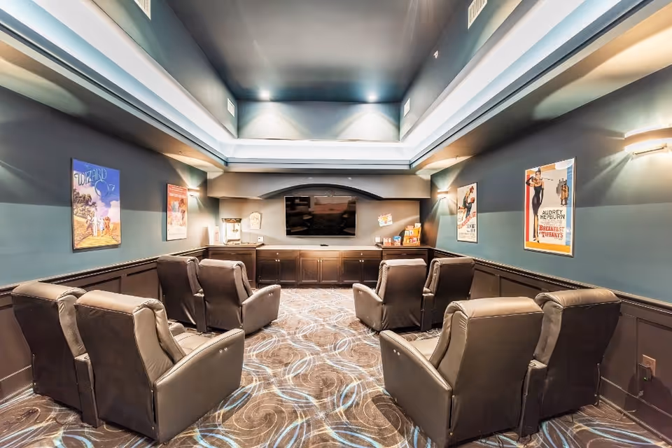 A cozy home theater room with eight black leather recliner chairs arranged in two rows facing a large flat-screen TV mounted on a dark wood cabinet. The walls are painted dark teal and decorated with classic movie posters including The Wizard of Oz and Breakfast at Tiffany's. The ceiling has recessed lighting and a tray design, and the carpet features a swirling pattern in shades of brown and blue.