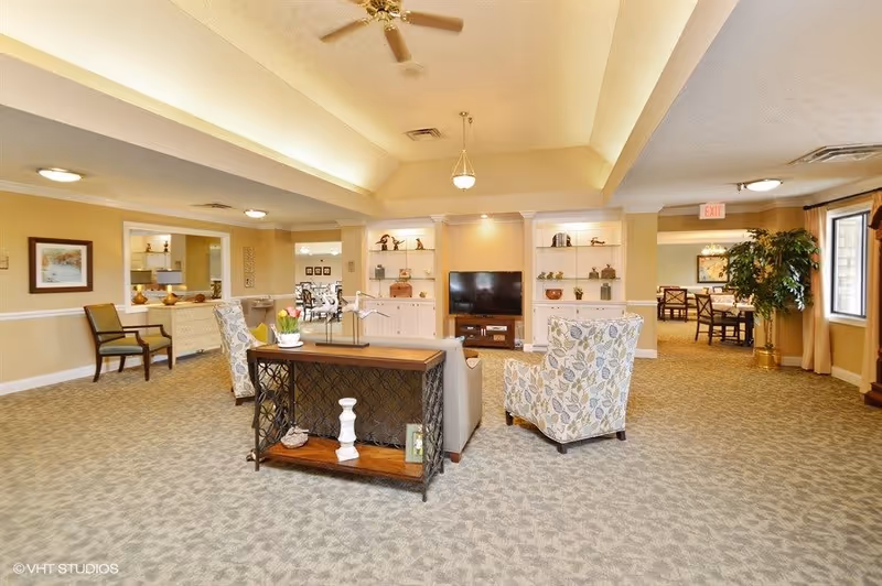 A spacious and well-lit senior living facility common area with patterned carpet, beige walls, and a high ceiling with ceiling fans and hanging lights. The room features a TV on a stand flanked by built-in shelves with decorative items, two floral-patterned armchairs, a small wooden table with decorative objects, and additional seating along the walls. In the background, there is a dining area with tables and chairs, large windows with curtains, and a potted plant near the window.