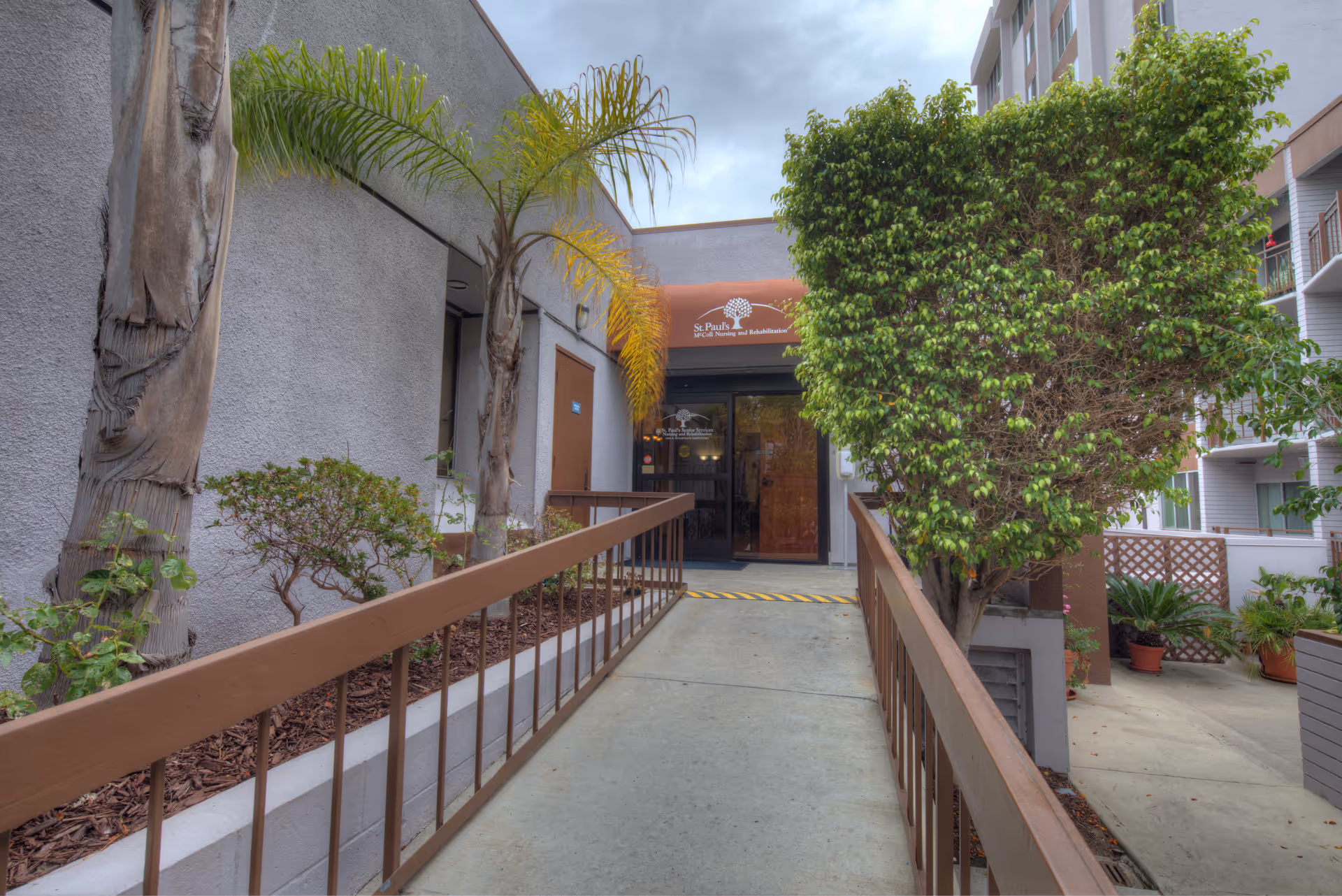 Entrance to St. Paul's Skilled Nursing and Rehabilitation facility with a concrete ramp flanked by brown railings, palm trees, and other greenery on either side. The building has a gray exterior, and there is an orange awning above the glass door entrance displaying the facility's name and logo.