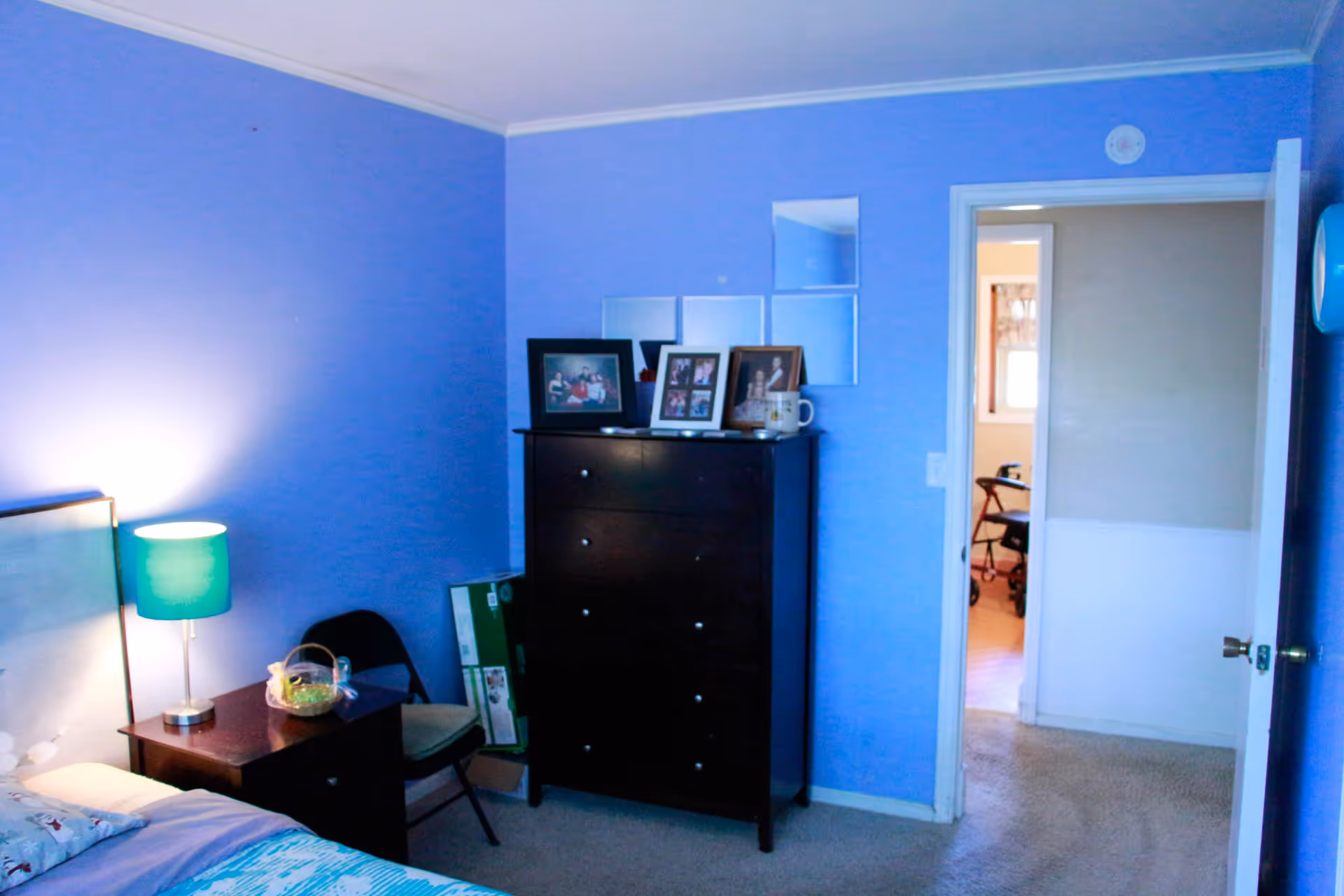 A bedroom with blue walls featuring a bed with a patterned bedspread, a nightstand with a green lamp and a small basket, a black chair, and a tall dark dresser with framed photos on top. The door is open, showing a glimpse of another room with a walker.