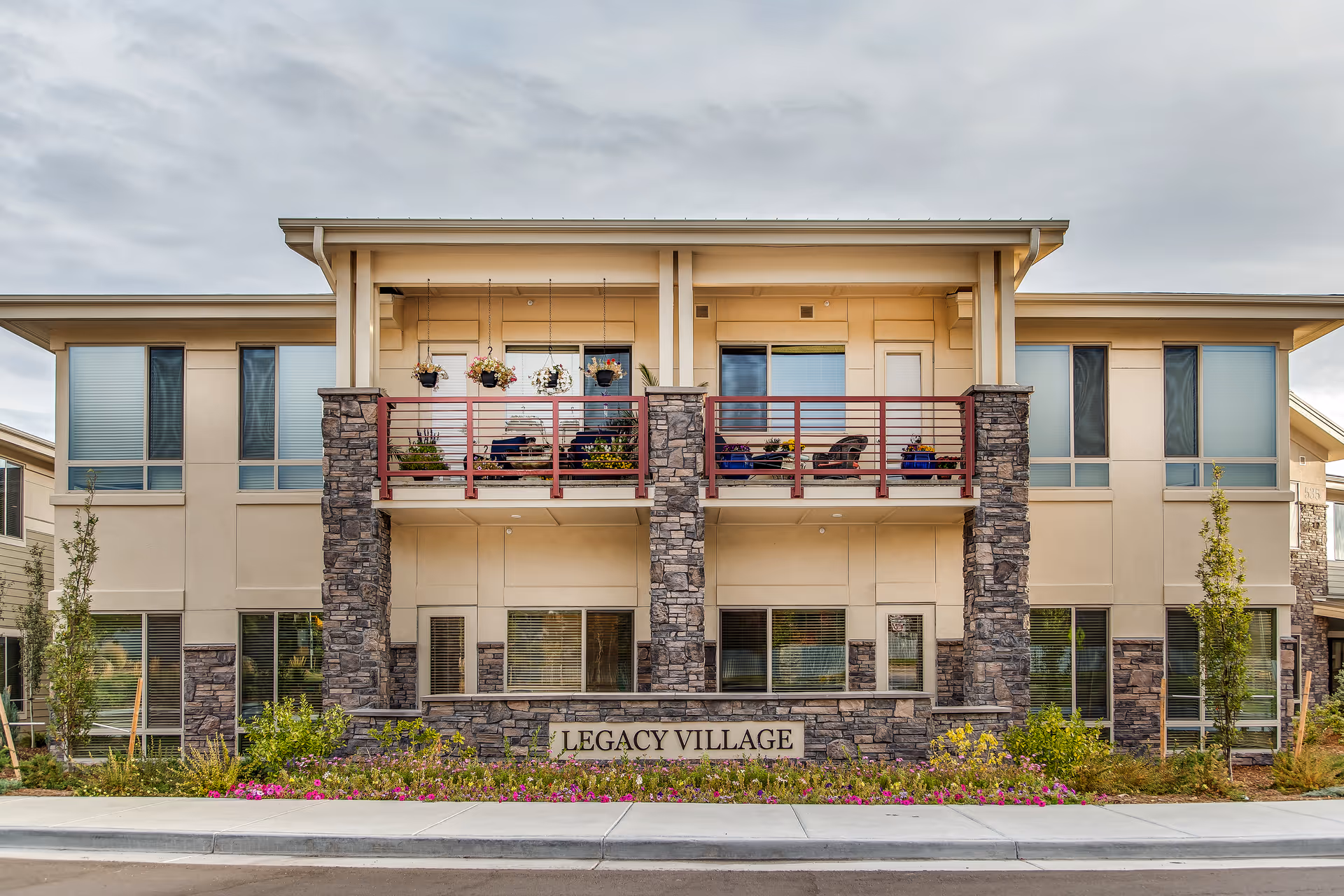 Front exterior of the Legacy Village building with balconies, stone pillars, landscaping, and a sign reading 'LEGACY VILLAGE'.