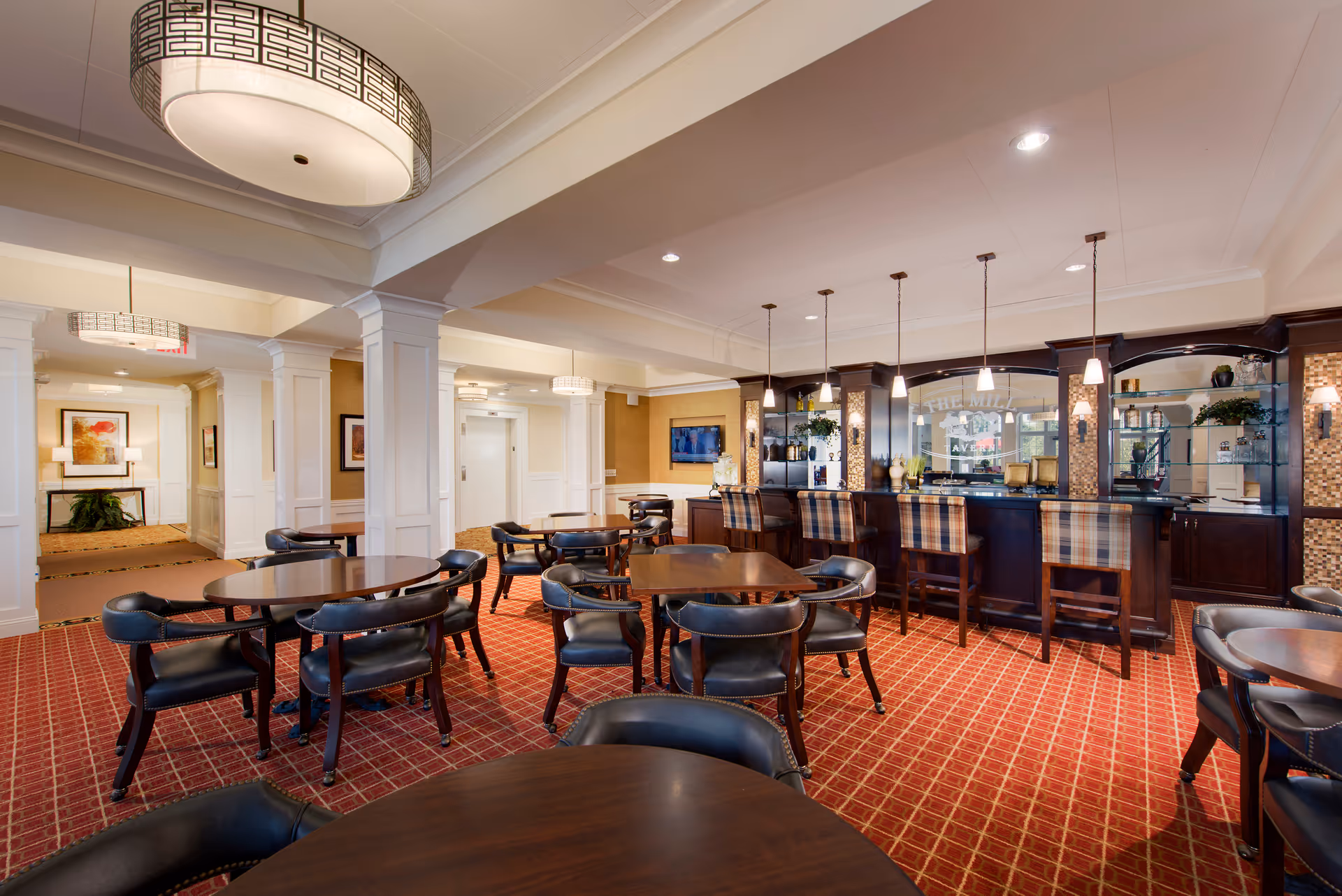 A spacious dining area with several round wooden tables and black cushioned chairs arranged on a red patterned carpet. The room features white columns and ceiling beams, with modern pendant lights hanging above a dark wood bar counter with plaid upholstered bar stools. The bar area has glass shelves with decorative items and a mirror behind it. The walls are painted in warm tones with framed artwork and a television mounted on one wall.