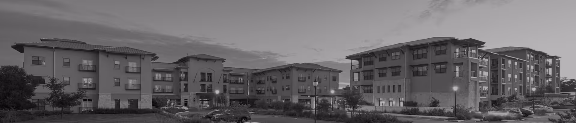 Panoramic exterior view of a multi-story assisted living building with landscaped grounds and parked cars at dusk.