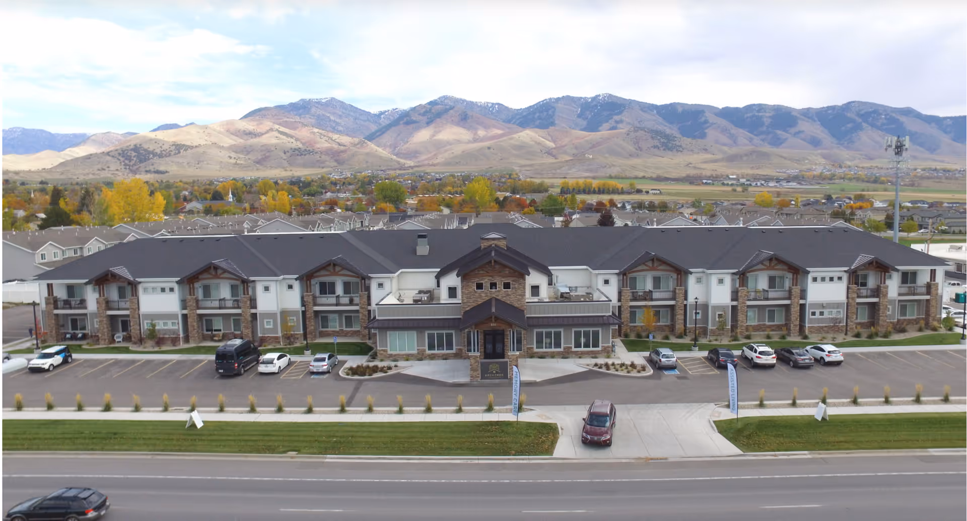 Front exterior view of Birch Creek Assisted Living of Smithfield, a two-story building with balconies, parking spaces, and a mountainous landscape in the background.