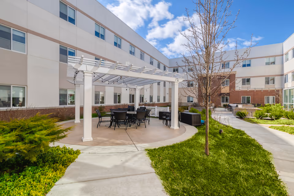 Courtyard with a white pergola over tables and chairs, walkways, grass, landscaping, and a surrounding multi-story building.