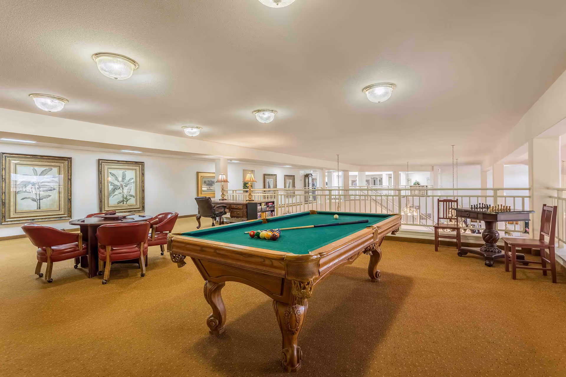 A spacious recreational room with a pool table in the center, a round table with four red leather chairs, and a small chess table with two wooden chairs. The room has beige carpet, white walls, framed botanical artwork, and multiple ceiling lights. There is a railing overlooking a lower level.
