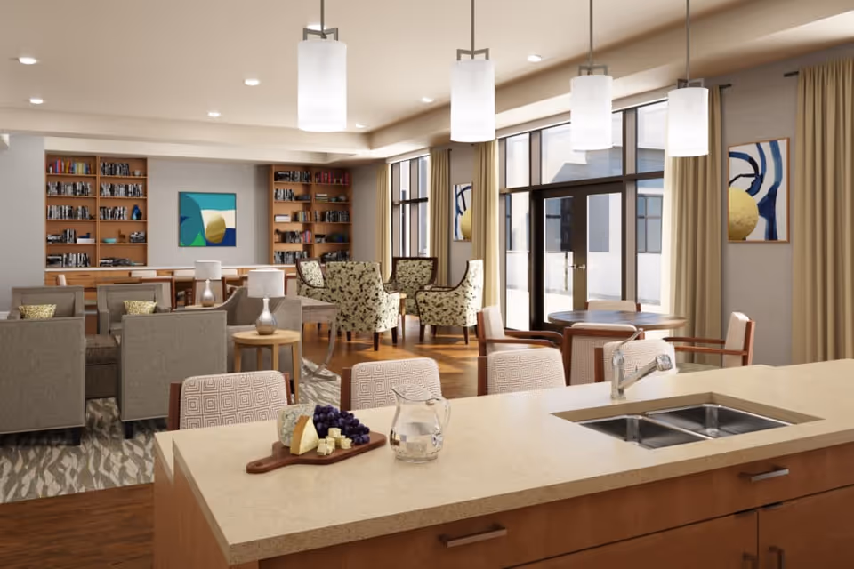 Open communal living area with a kitchen island in the foreground, multiple seating groups, bookshelves and large windows.