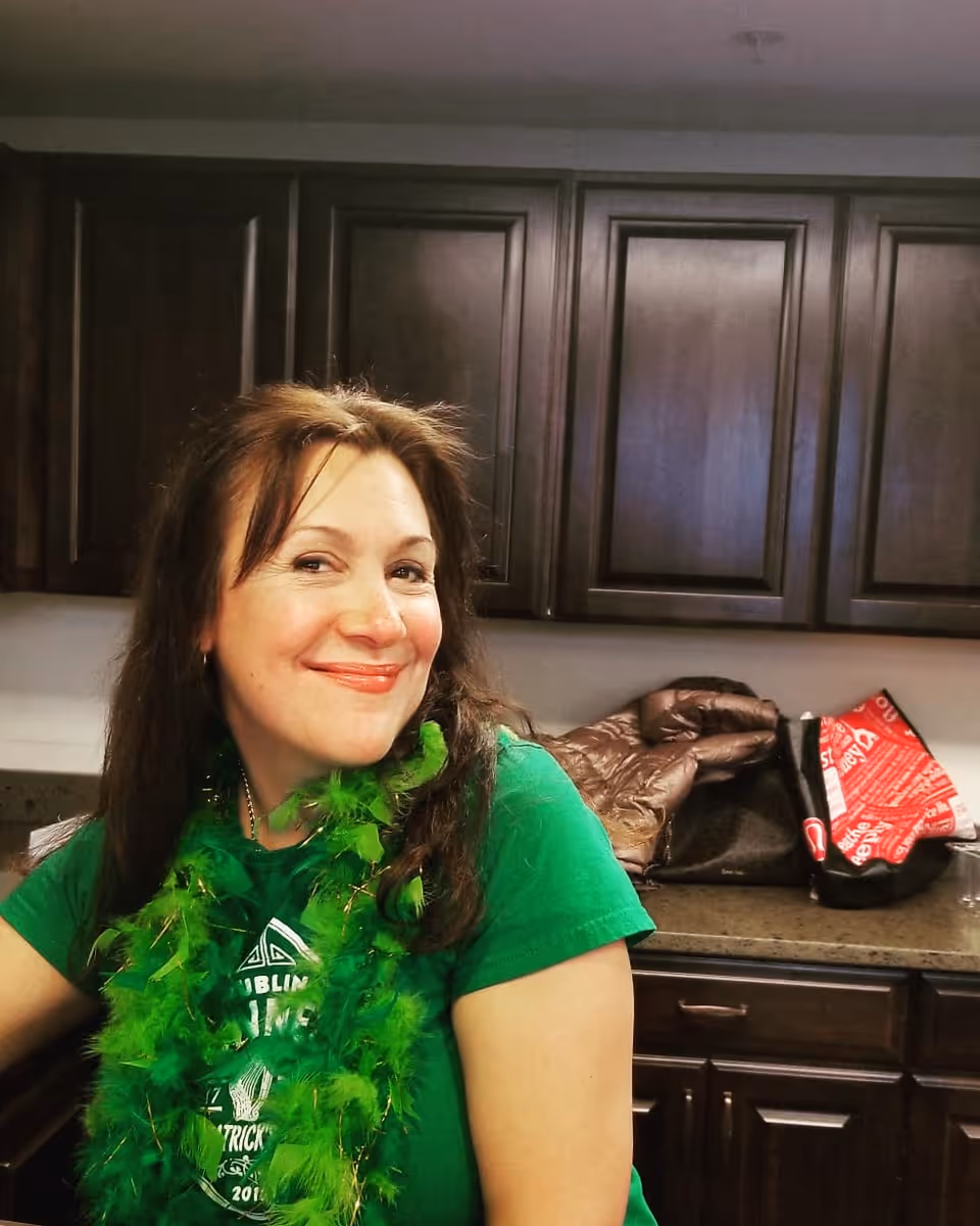 A woman with long brown hair wearing a green shirt and a green feather boa is smiling while standing in a kitchen with dark wooden cabinets and a countertop with bags and a jacket on it.
