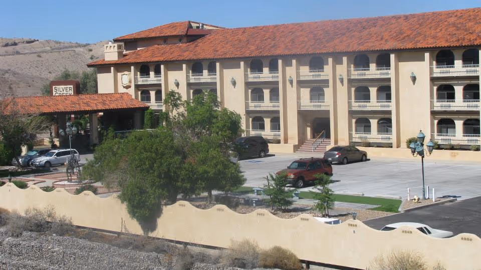Exterior view of Silver Creek Assisted Living facility showing a multi-story building with balconies, a covered entrance, several parked cars, trees, and a beige decorative wall in the foreground under a clear blue sky.