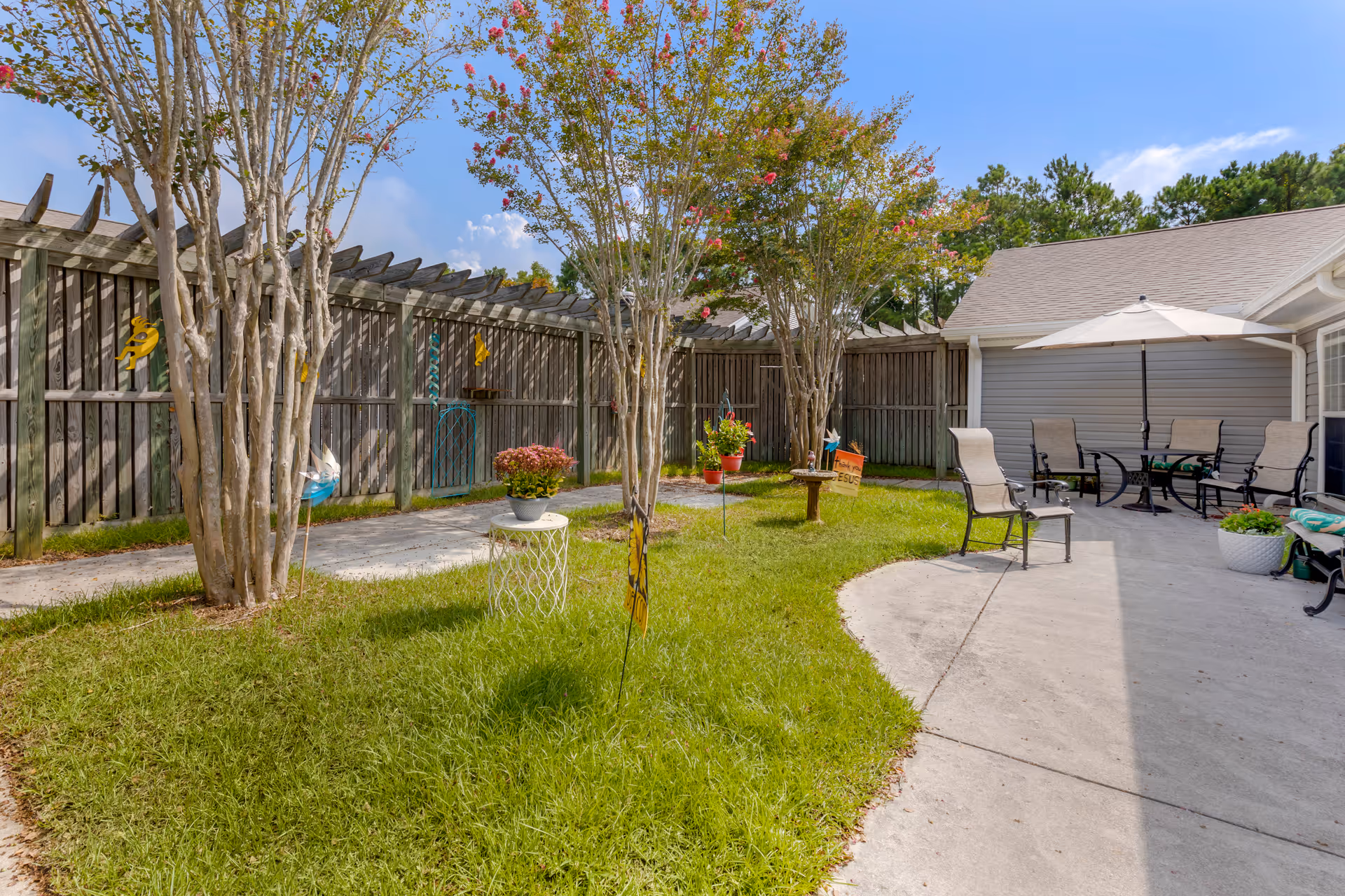 Fenced sunlit courtyard with small trees, grass, potted plants, and patio chairs with an umbrella next to a concrete walkway.