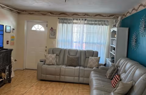 A living room with two gray leather sofas arranged around a window with sheer curtains. There is a white door to the left, a dark wooden TV stand with a television, and a white bookshelf on the right against a teal accent wall decorated with metallic wall art.