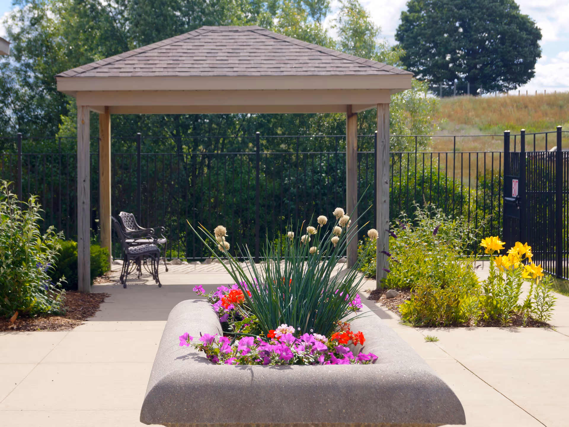 Outdoor garden area with a concrete planter filled with colorful flowers and tall grass. In the background, there is a wooden gazebo with a shingled roof and two metal chairs underneath. The area is surrounded by greenery and a black metal fence.