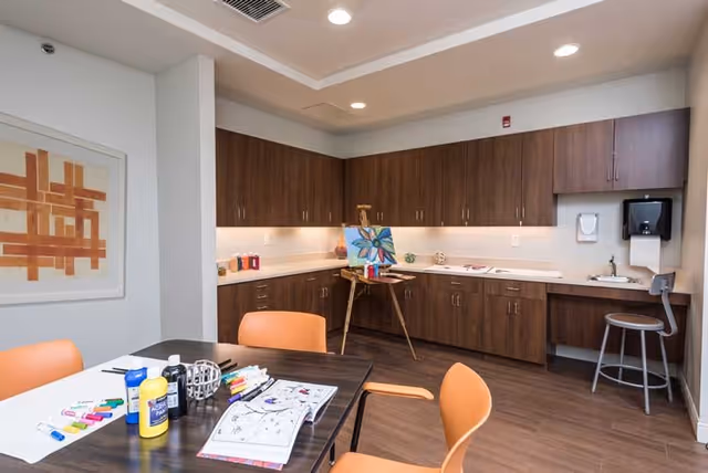 A well-lit room with wooden cabinets along the back wall, a countertop with art supplies, and an easel holding a painting of a blue flower. In the foreground, there is a table with orange chairs around it, featuring various art materials including paint bottles, markers, and coloring books. The room has a modern ceiling with recessed lighting and a framed abstract artwork on the left wall.