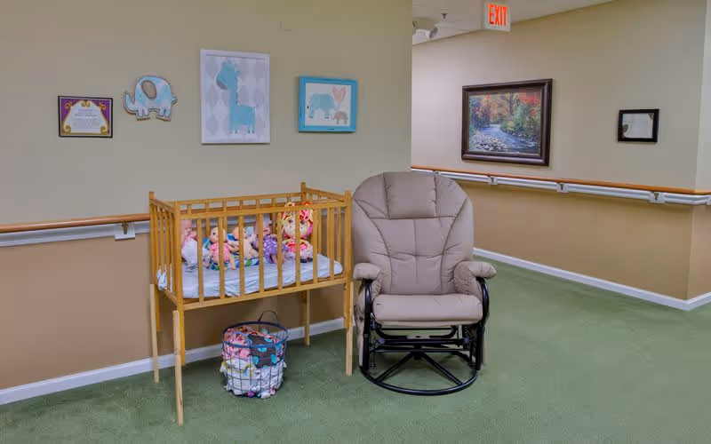 A hallway corner in a senior living facility with a wooden crib containing stuffed animals and dolls, a beige cushioned rocking chair, a basket with blankets, and framed pictures on the walls including animal illustrations and a landscape painting.