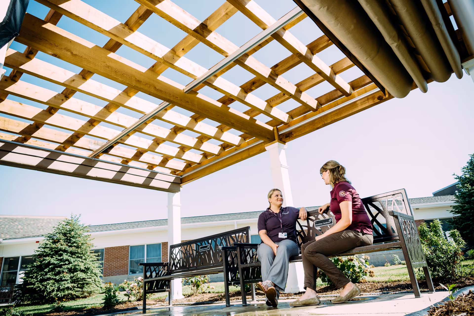 Two women sitting and talking on metal benches under a wooden pergola outside a building with a garden and trees in the background on a sunny day.