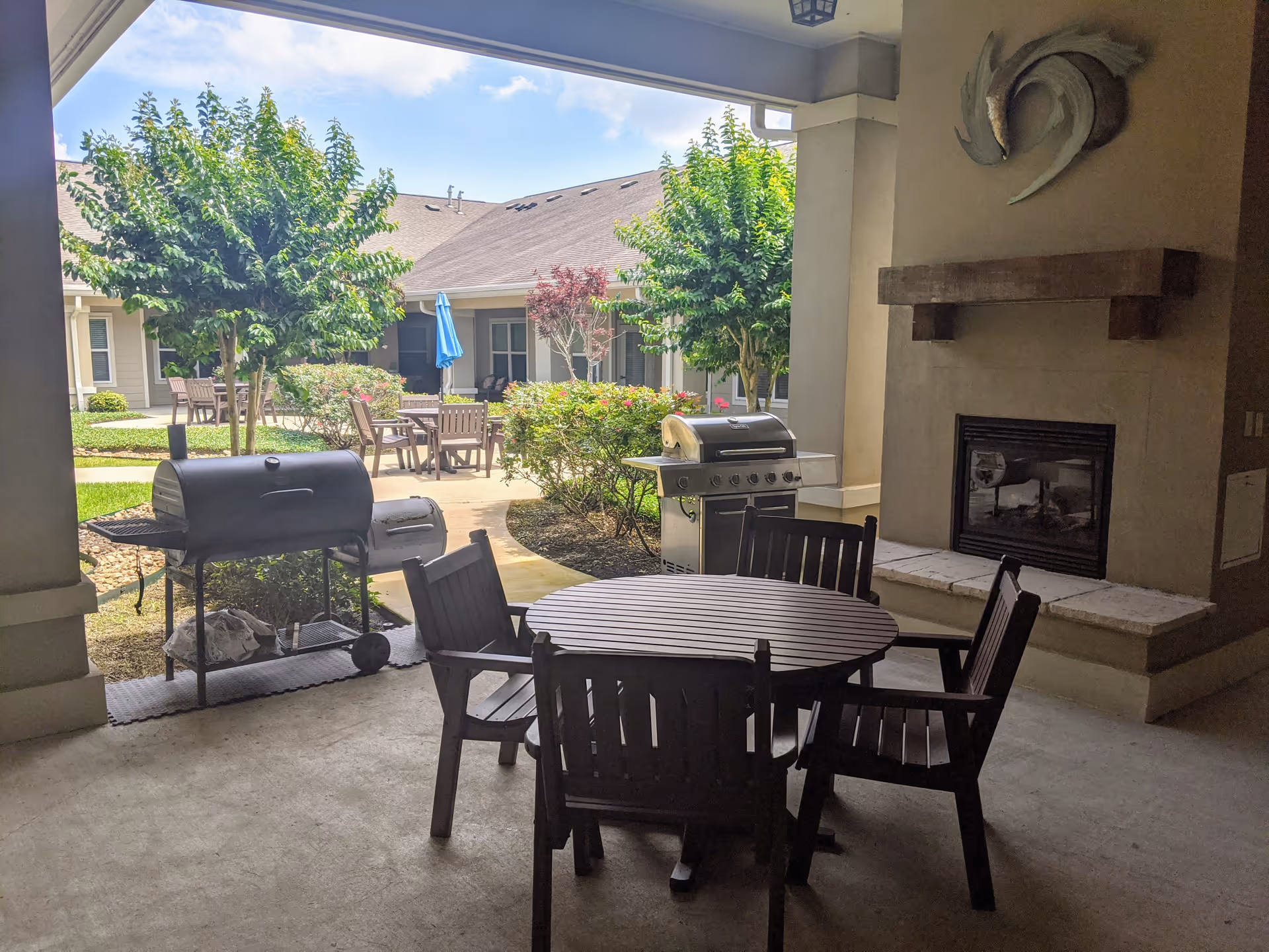Covered outdoor patio area with a round wooden table and four chairs, two grills, a fireplace with a decorative sculpture above it, and a view of a courtyard with trees, bushes, and additional seating under a blue umbrella.