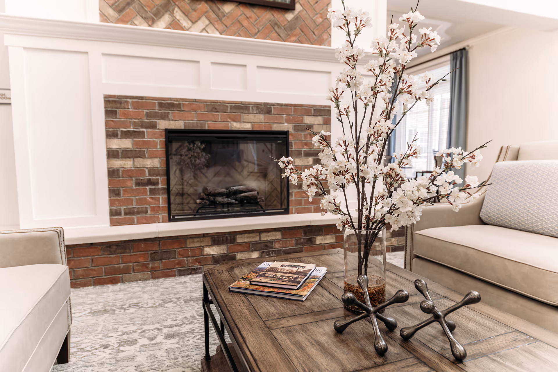 A cozy living room with a brick fireplace, beige sofas, a wooden coffee table with decorative items including a vase with white flowers and two metal jack sculptures, and a patterned carpet.