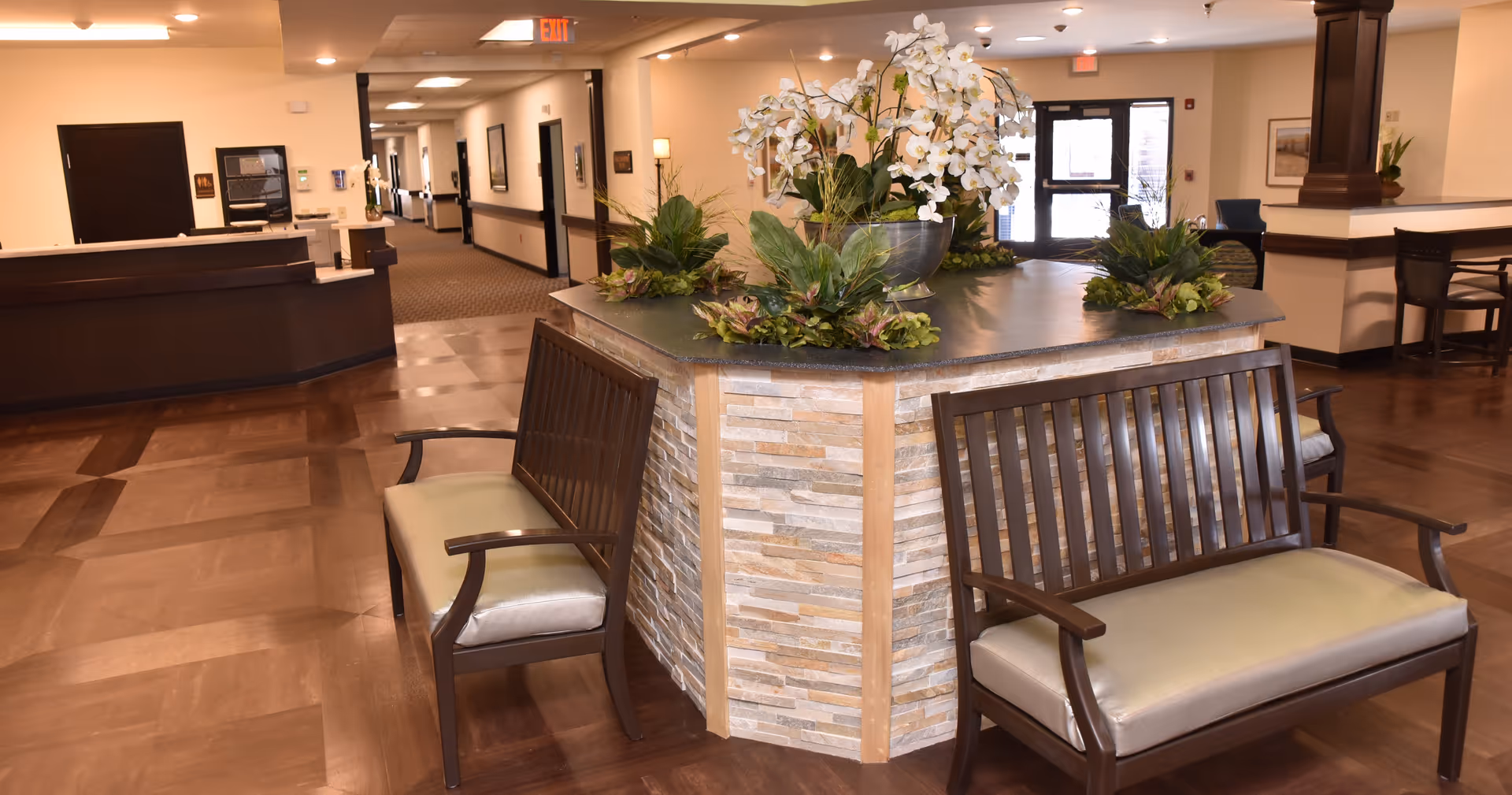 Interior view of a senior living facility lobby area with a central stone planter decorated with white orchids and green plants. Surrounding the planter are wooden benches with beige cushions. The floor is polished wood, and there is a reception desk to the left. The hallway extends into the background with doors and framed pictures on the walls. The entrance doors are visible in the background.