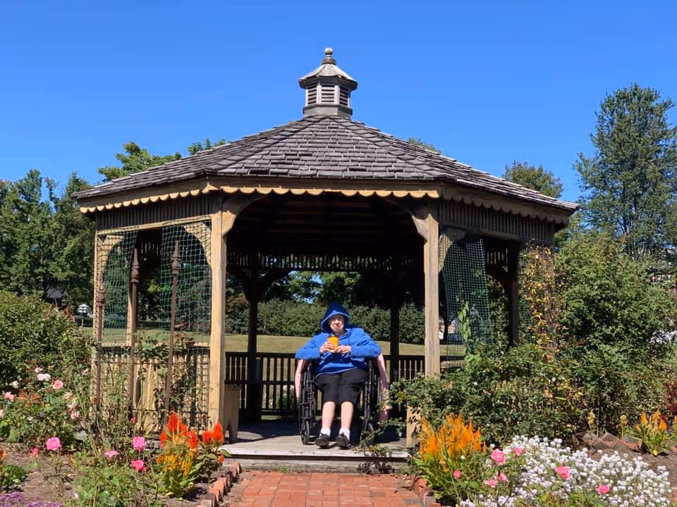 A person in a blue hoodie sitting in a wheelchair inside a wooden gazebo surrounded by colorful flowers and greenery under a clear blue sky.