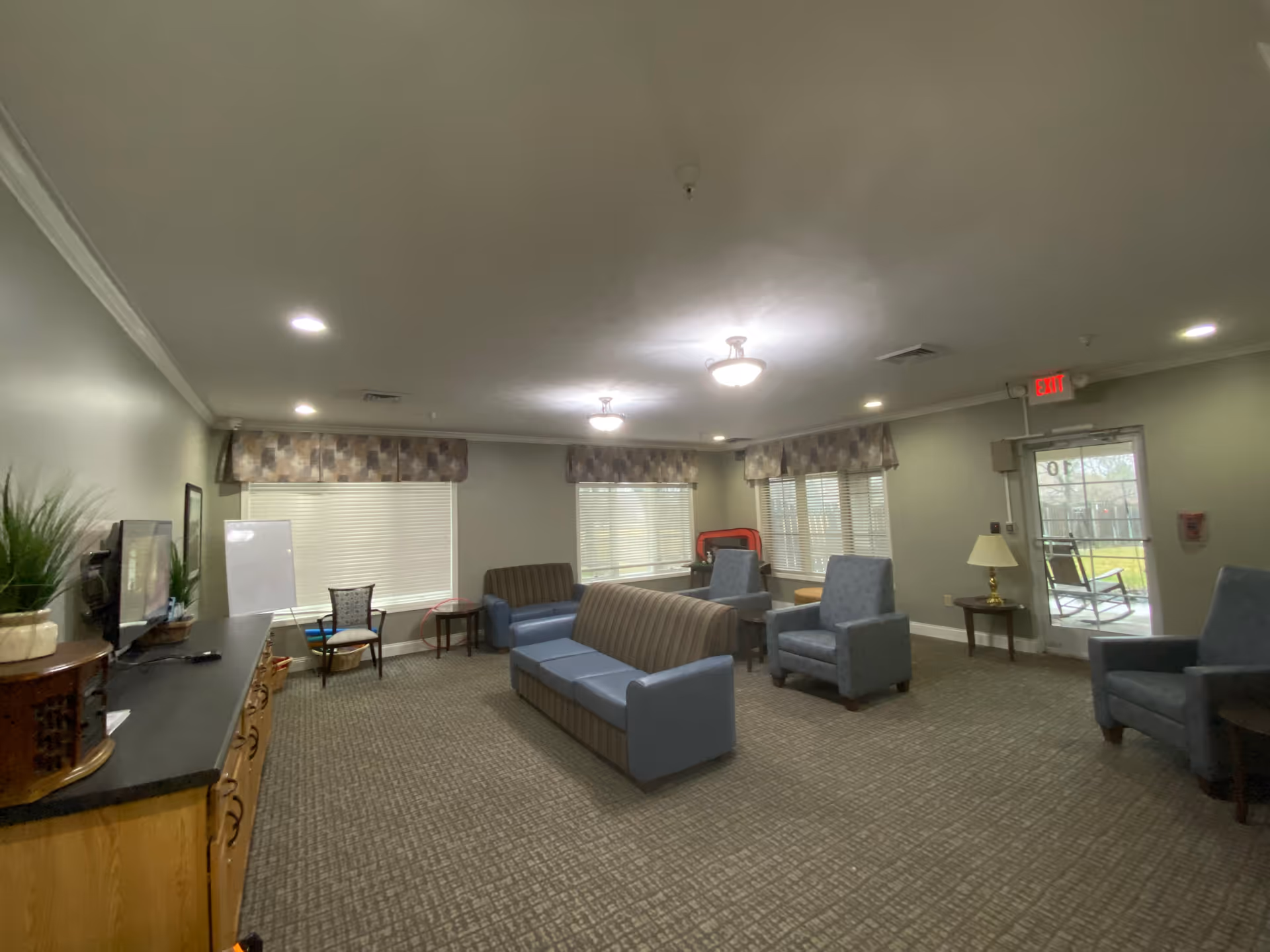 A spacious common living area with multiple seating options including two blue armchairs, two striped sofas, and a wooden chair. The room has carpeted flooring, beige walls, and several windows with patterned valances allowing natural light. There is a wooden cabinet with a TV on top, a small table with a lamp near a glass door leading outside, and ceiling lights providing illumination.