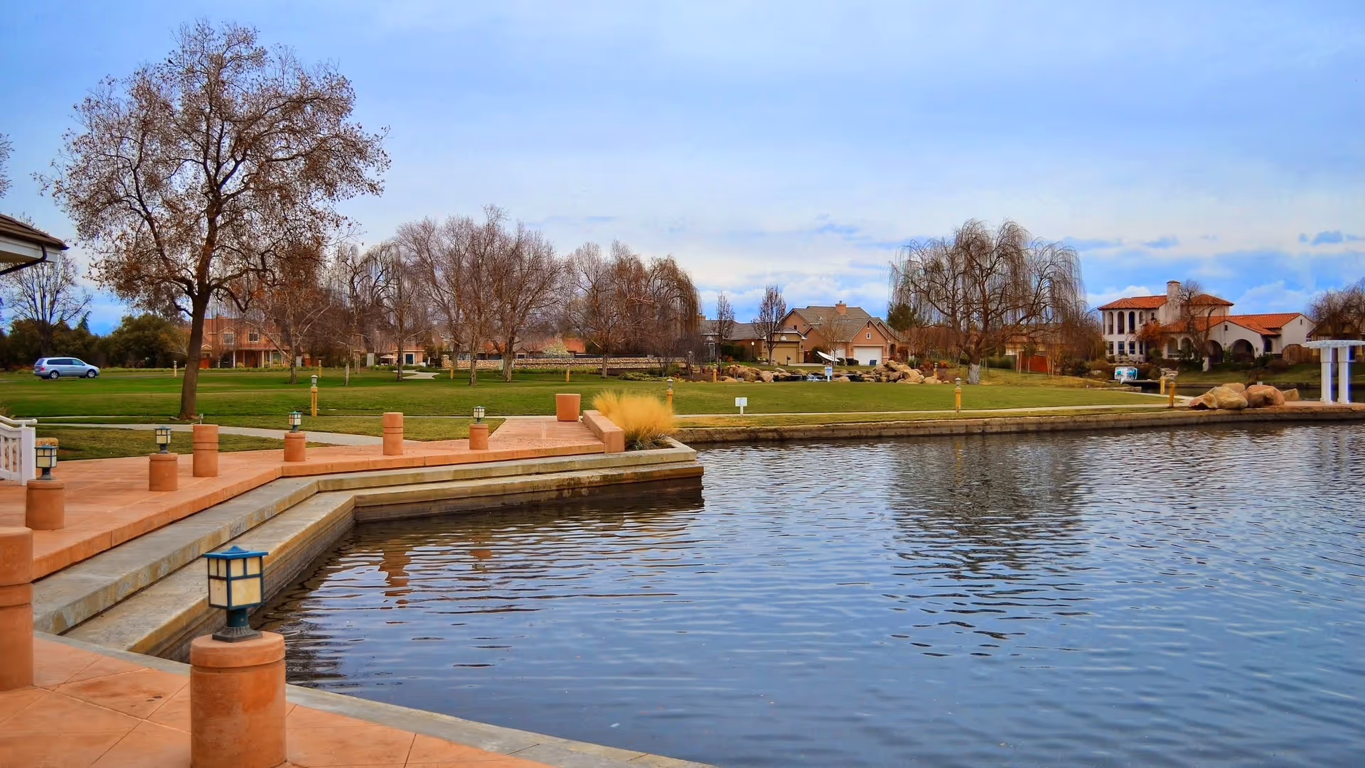 A serene outdoor scene featuring a large pond with calm water, surrounded by a paved walkway with small lantern-style lights. In the background, there is a grassy area with leafless trees and several houses under a partly cloudy sky.