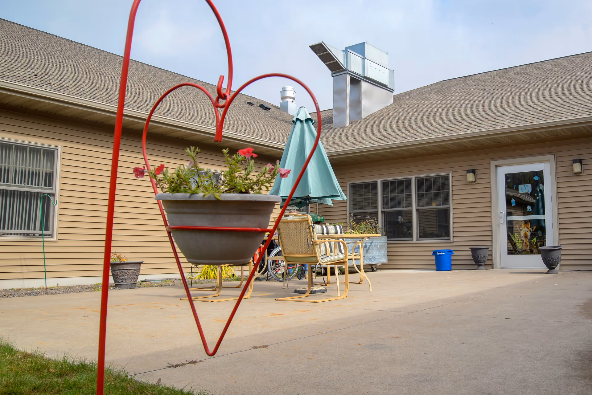 Outdoor patio area at VitaCare Living of Proctor with a concrete floor, beige siding building, a heart-shaped red metal plant holder with flowers, patio chairs, a table with a closed green umbrella, and a door with windows.