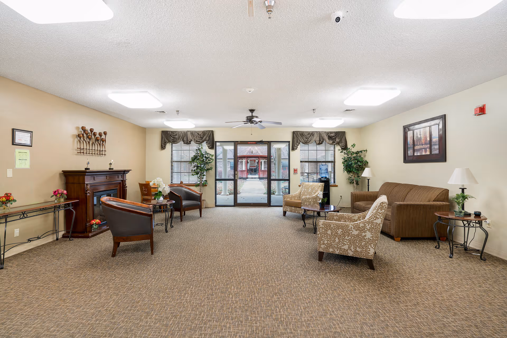 A spacious senior living facility common area with beige walls and carpeted floor. The room features a brown sofa, patterned armchairs, small tables with lamps and plants, a decorative fireplace, and large windows with valances. Double glass doors lead outside, and ceiling lights and a fan provide illumination.