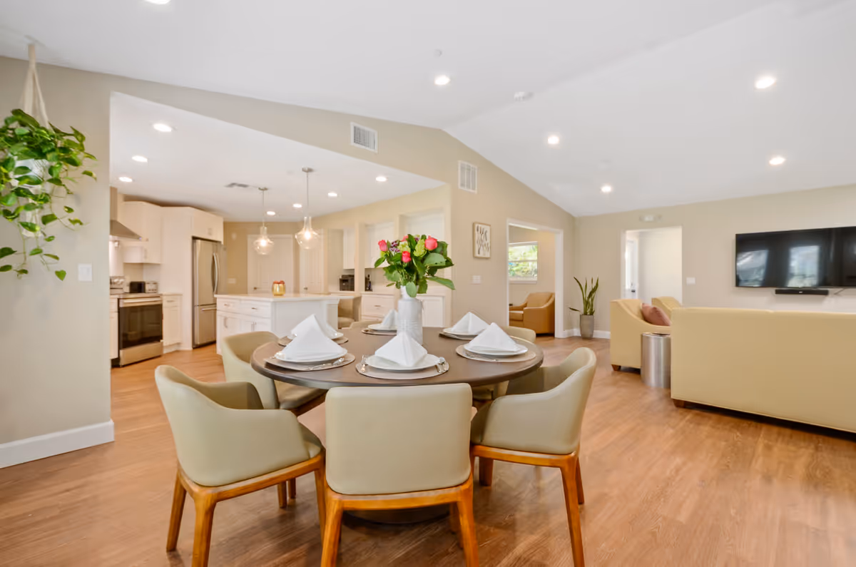 A bright and spacious open-concept living area featuring a round dining table set with six chairs and neatly folded white napkins on plates. A vase with colorful flowers is centered on the table. The background shows a modern kitchen with white cabinetry, stainless steel appliances, and pendant lights. Adjacent to the dining area is a living room with beige sofas, a wall-mounted flat-screen TV, and potted plants, all set on wood flooring under recessed ceiling lights.