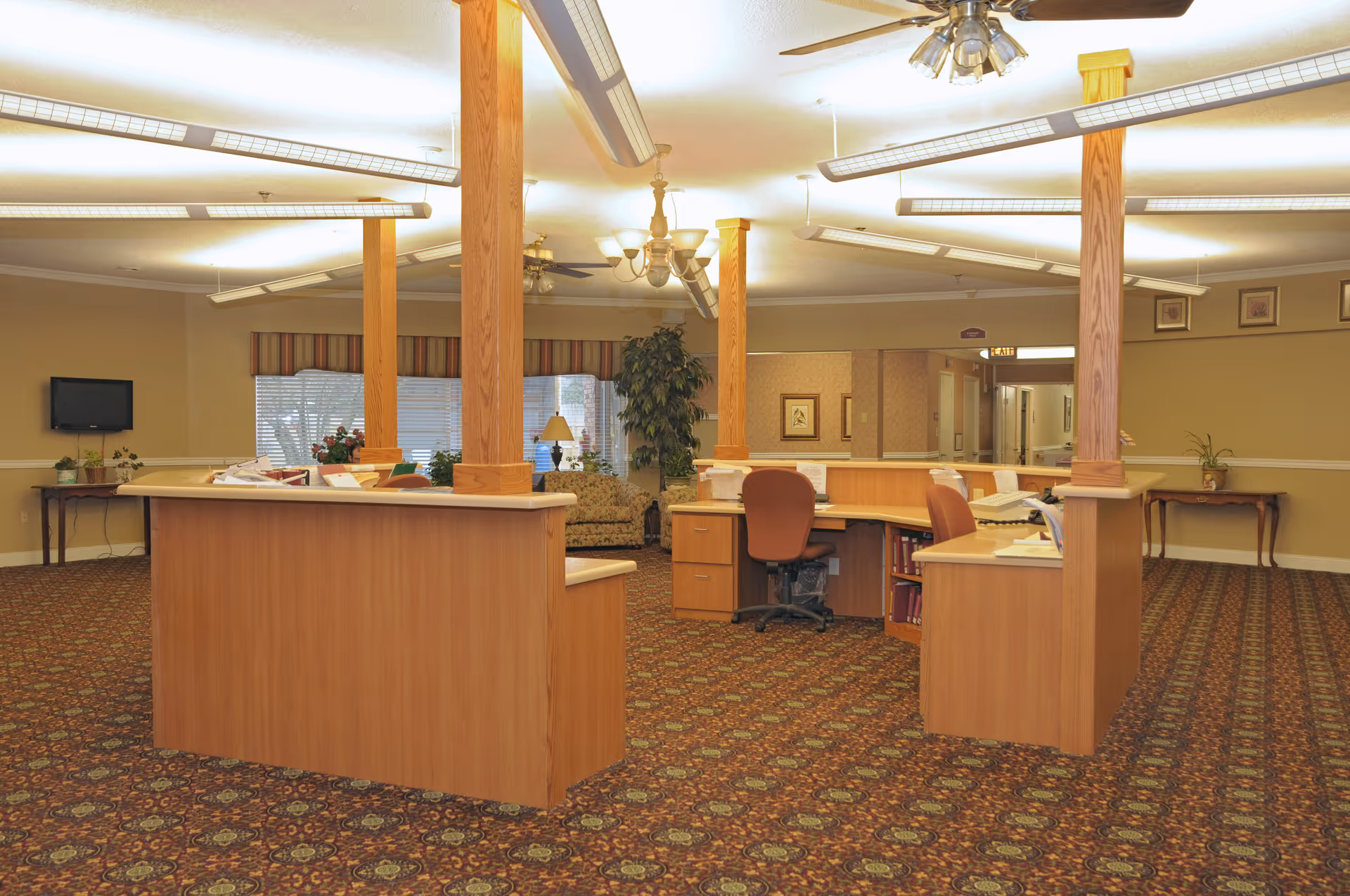 Reception area in a senior living facility with wooden desks, office chairs, carpeted floor with patterned design, ceiling fans, fluorescent lighting, and a seating area with a sofa and plants near windows with striped valances.