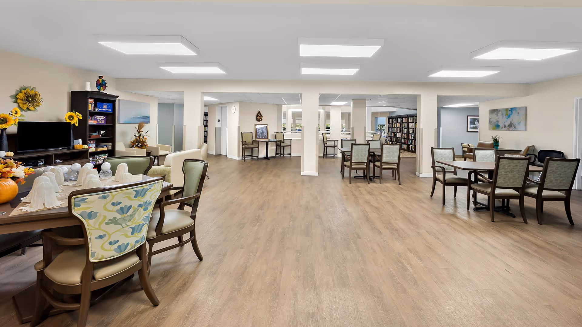 A spacious, well-lit common area in a senior living facility with multiple tables and chairs arranged for socializing or activities. The room features wood flooring, neutral-colored walls, and ceiling lights. There is a TV on a stand with shelves holding decorative items and books, along with some seasonal decorations like pumpkins and sunflowers.
