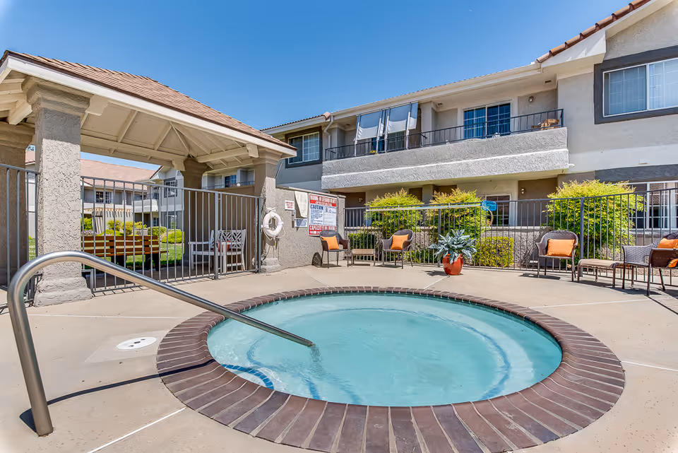 Outdoor hot tub with clear blue water surrounded by a concrete deck and metal railing. Several chairs with orange cushions and potted plants are arranged around the area. The background shows a two-story building with balconies and windows under a clear blue sky.