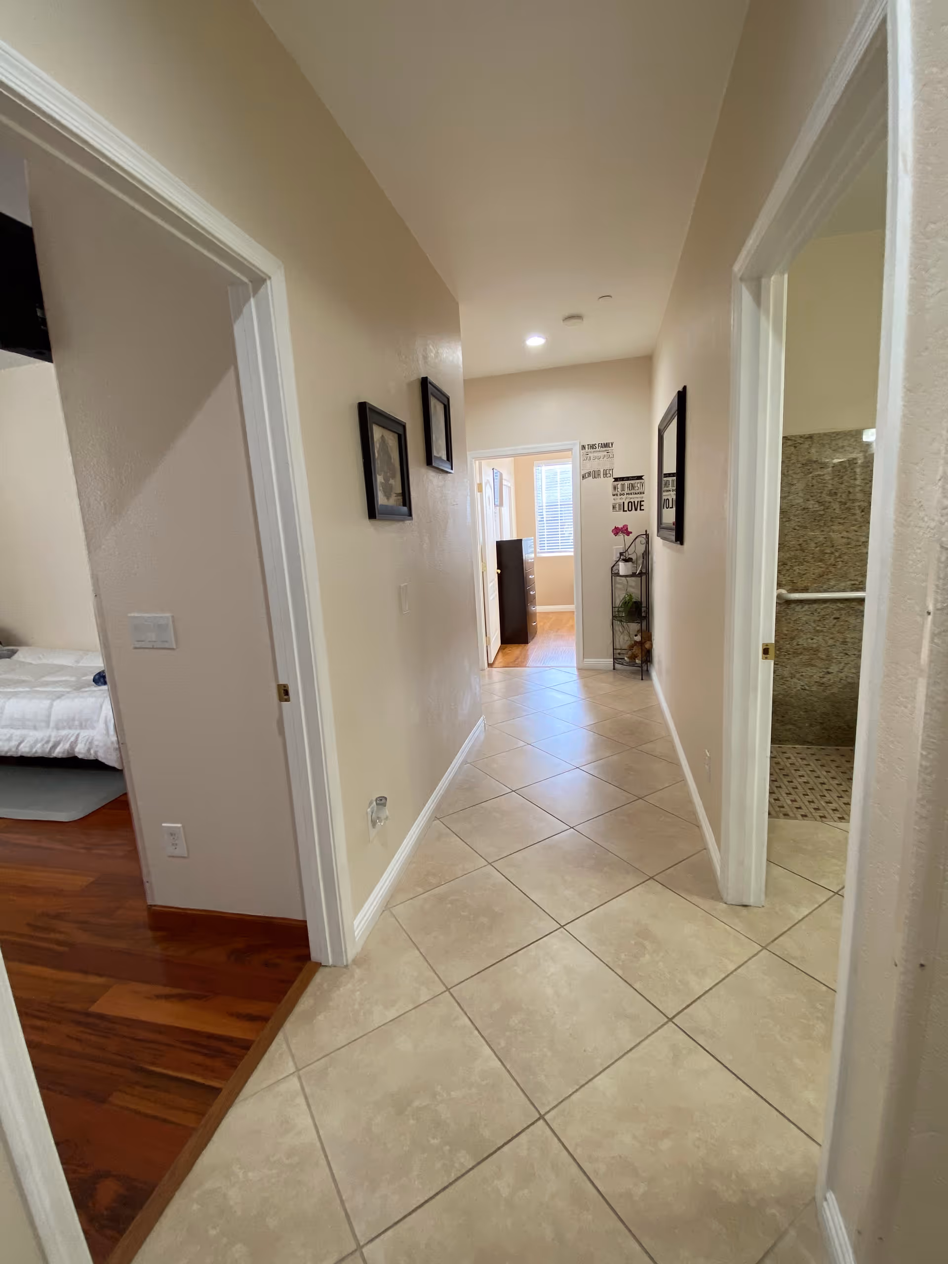 Tiled interior hallway with open bedroom and bathroom doors, framed pictures on the walls, and a small shelving unit at the far end.