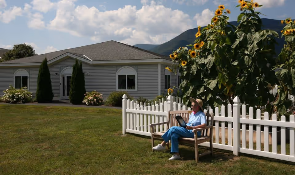 An elderly person wearing a hat and blue clothing sits on a wooden bench in a grassy yard next to a white picket fence with tall sunflowers behind it. In the background, there is a single-story building with gray siding, white trim, and two tall evergreen shrubs flanking the entrance. Mountains and a partly cloudy sky are visible in the distance.