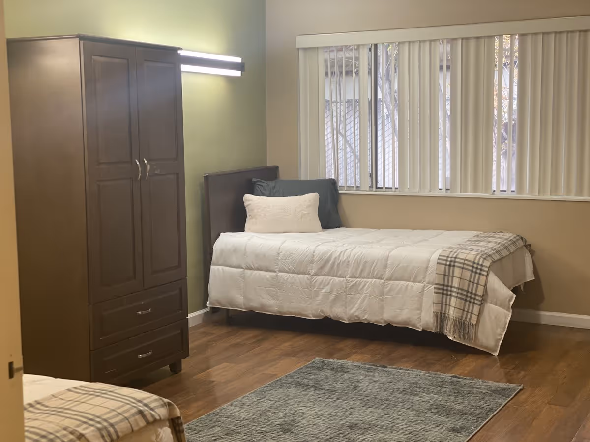 A simple bedroom with a single bed dressed in a white comforter and plaid throw, a wooden wardrobe, vertical blinds over a window, and a rug on hardwood floors.