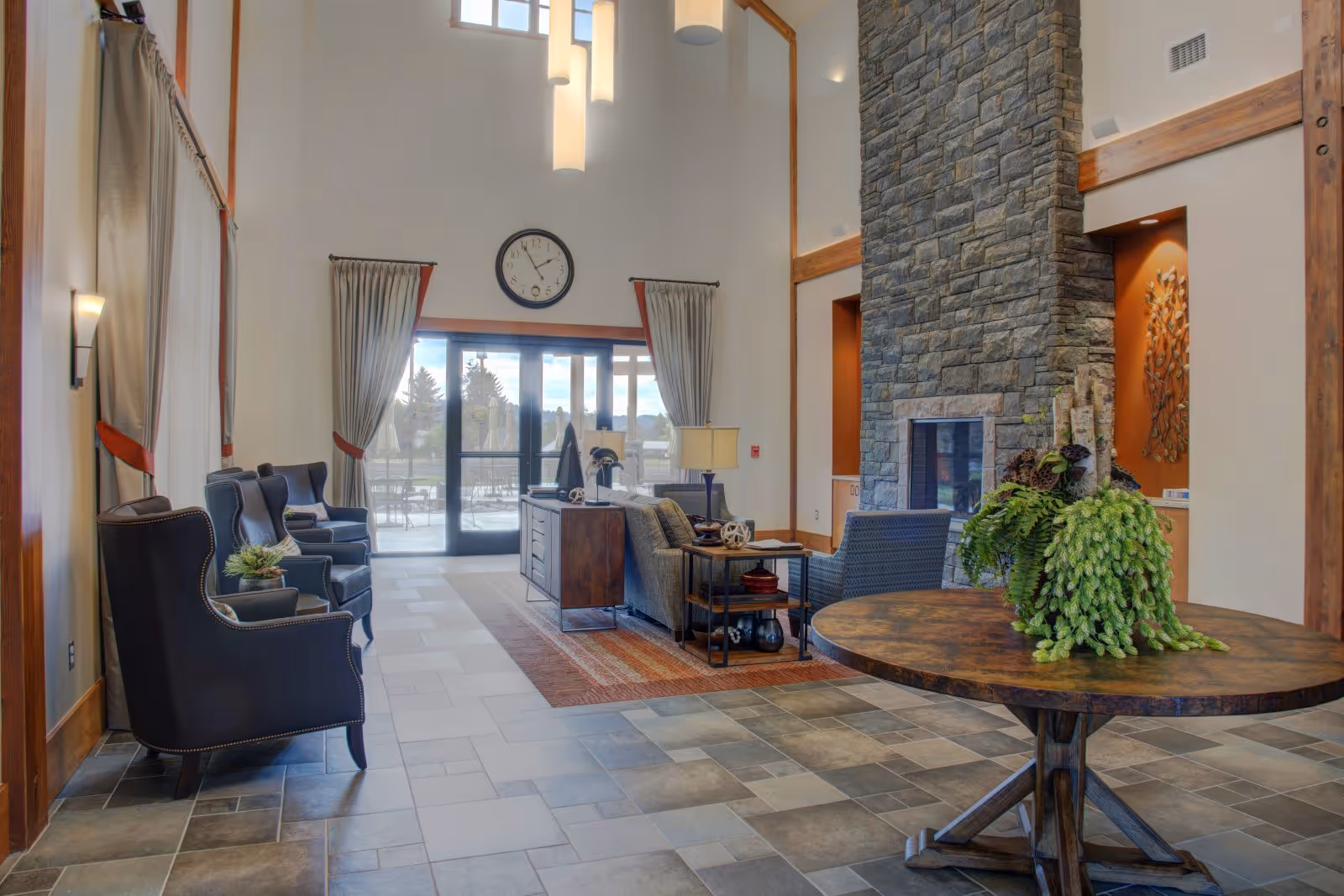 A spacious and well-lit common area with a high ceiling, featuring a stone fireplace, comfortable seating including armchairs and sofas, a round wooden table with a green plant centerpiece, large windows with curtains, and a wall clock above the glass doors leading outside.