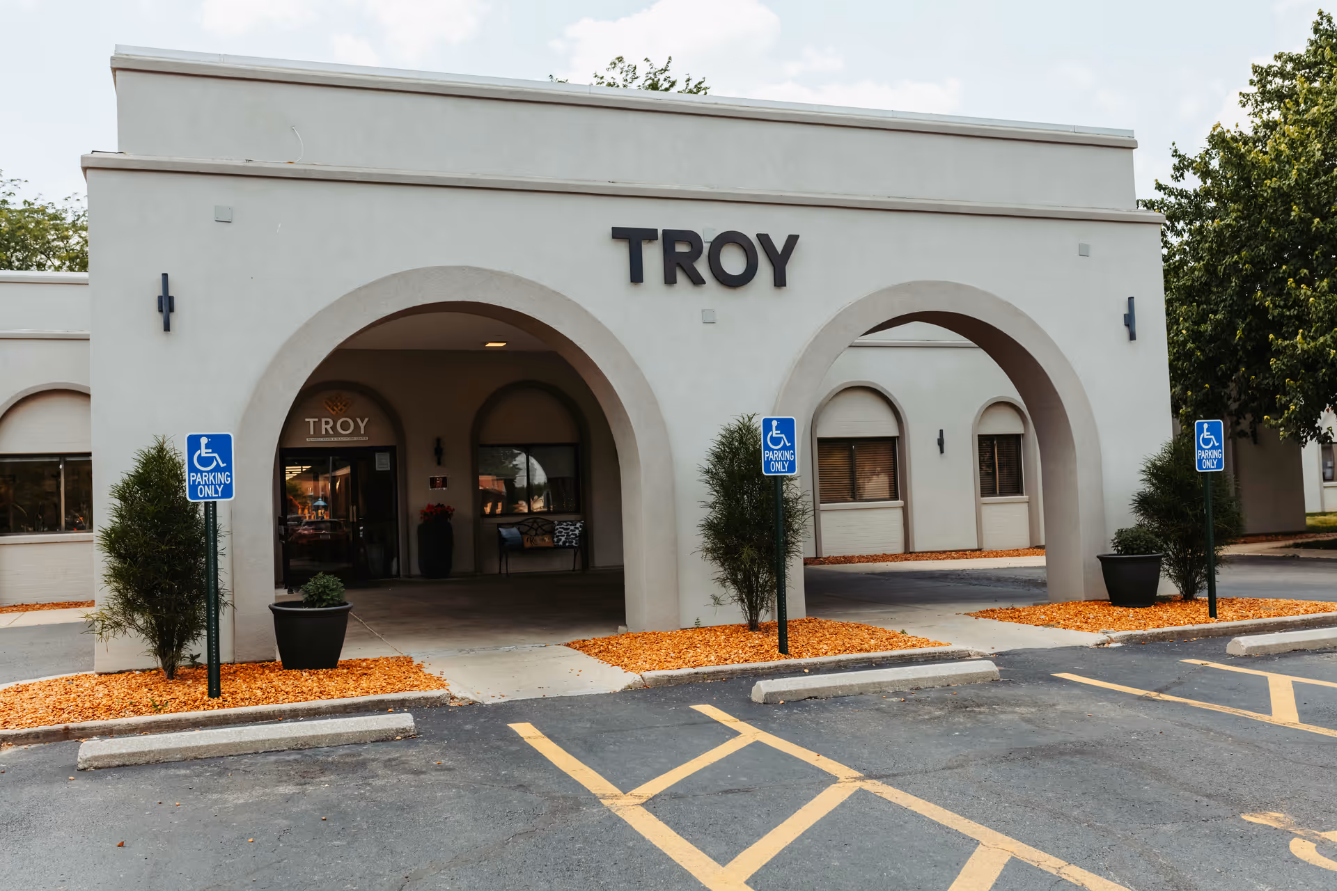 Front entrance of the Troy rehabilitation center with arched portico, large 'TROY' sign above, accessible parking signs and potted plants.