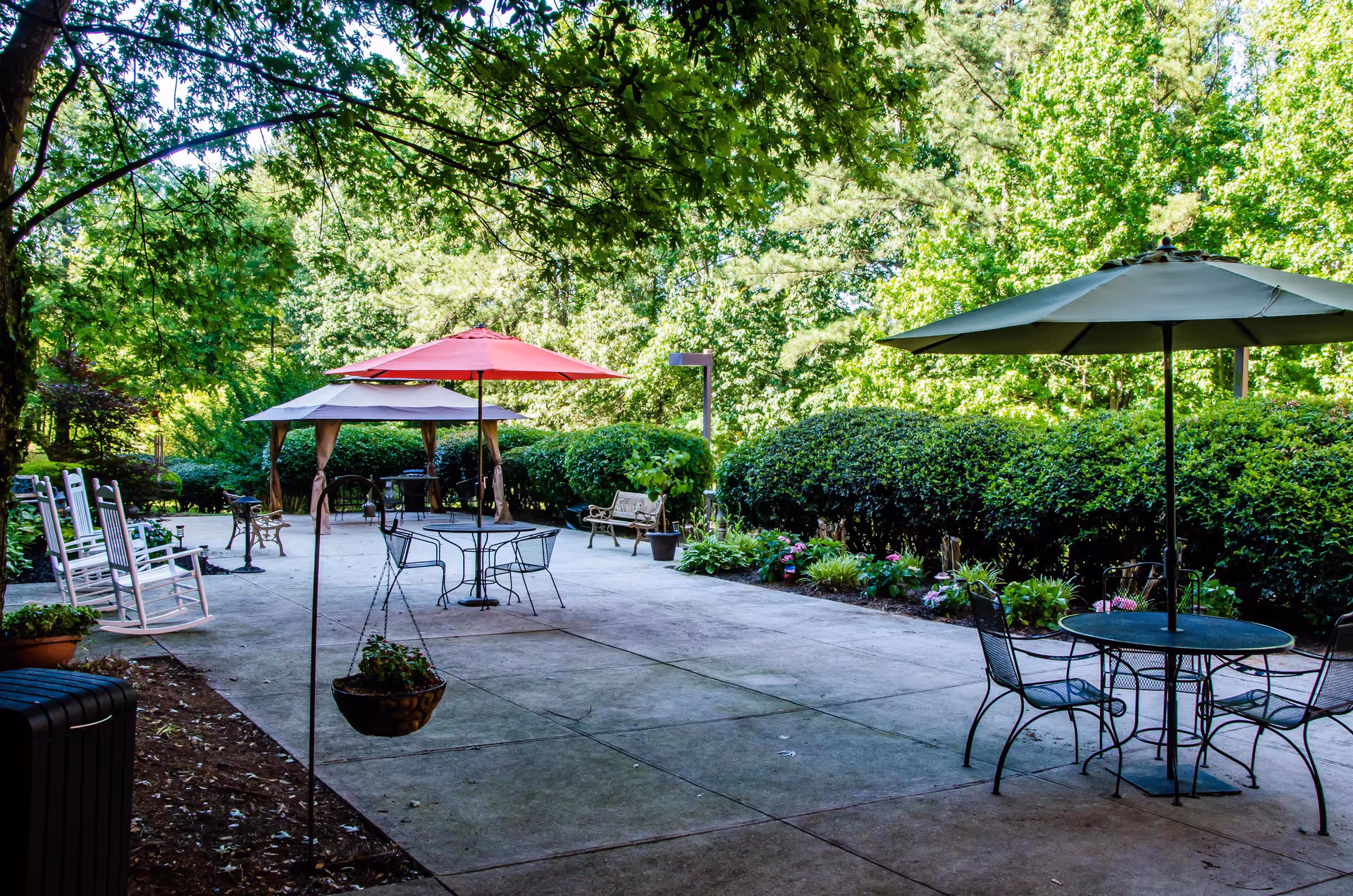 Outdoor patio area at Coventry Place with metal tables and chairs under umbrellas, white rocking chairs, benches, hanging flower pots, and lush green trees and bushes surrounding the space.