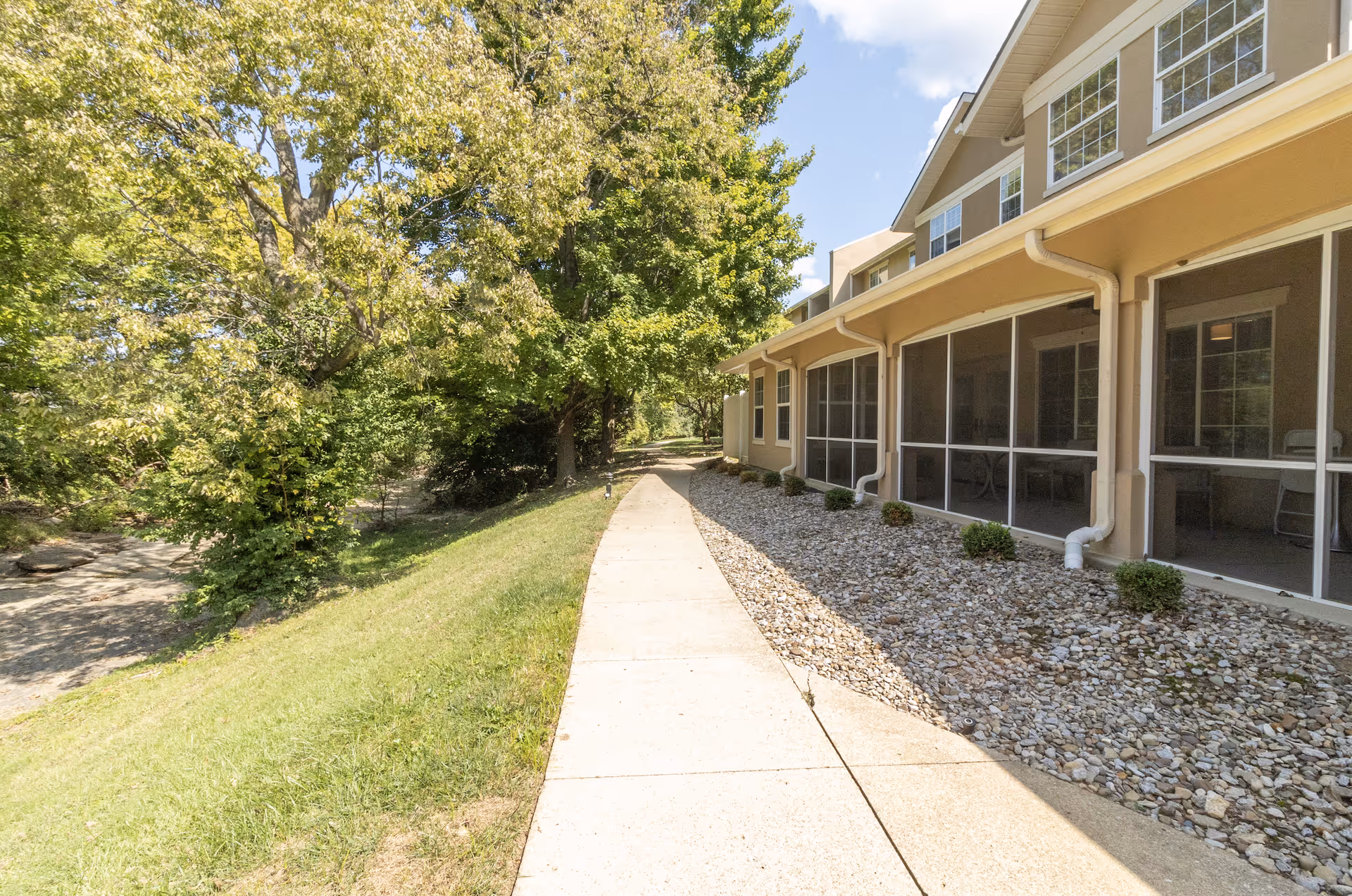 Side view of a building with screened-in porch areas along a paved walkway, bordered by a grassy slope and trees on one side under a partly cloudy sky.