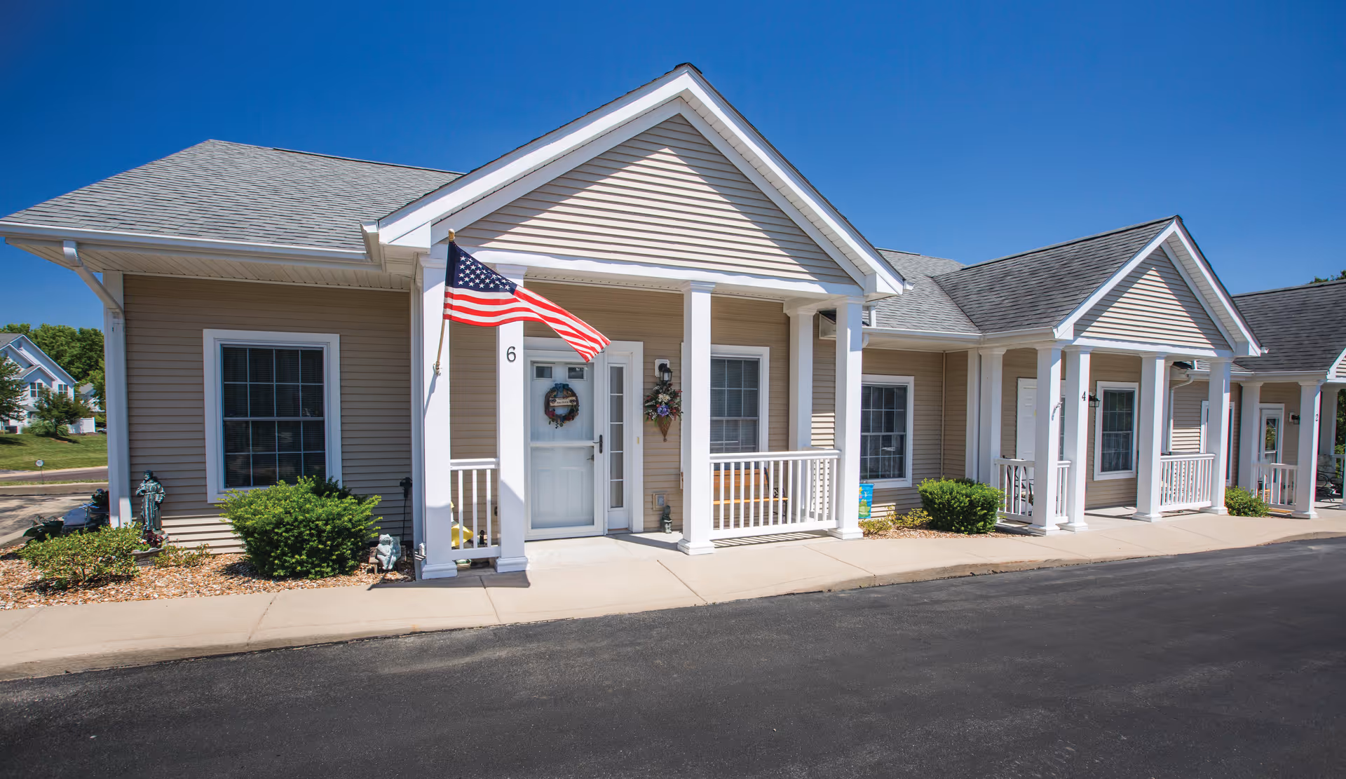 Exterior view of a single-story senior living facility building with beige siding, white trim, and a gray shingled roof. The building features small covered porches with white railings and columns. An American flag is mounted near the entrance door labeled with the number 6. There are bushes and small plants along the front of the building, and the sky is clear and blue.