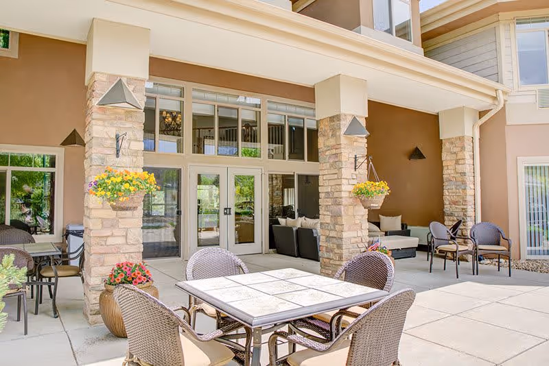 Outdoor patio area at a senior living facility with a tiled table and four wicker chairs in the foreground. Stone pillars with hanging flower baskets support a covered walkway leading to glass doors. Additional seating with cushioned chairs is visible under the covered area.