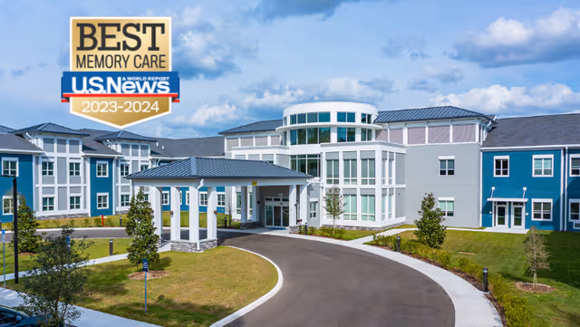 Exterior view of a modern senior living facility with a circular driveway, green lawns, and a clear blue sky. The building features large windows and a covered entrance. A badge in the top left corner reads 'Best Memory Care U.S. News 2023-2024'.