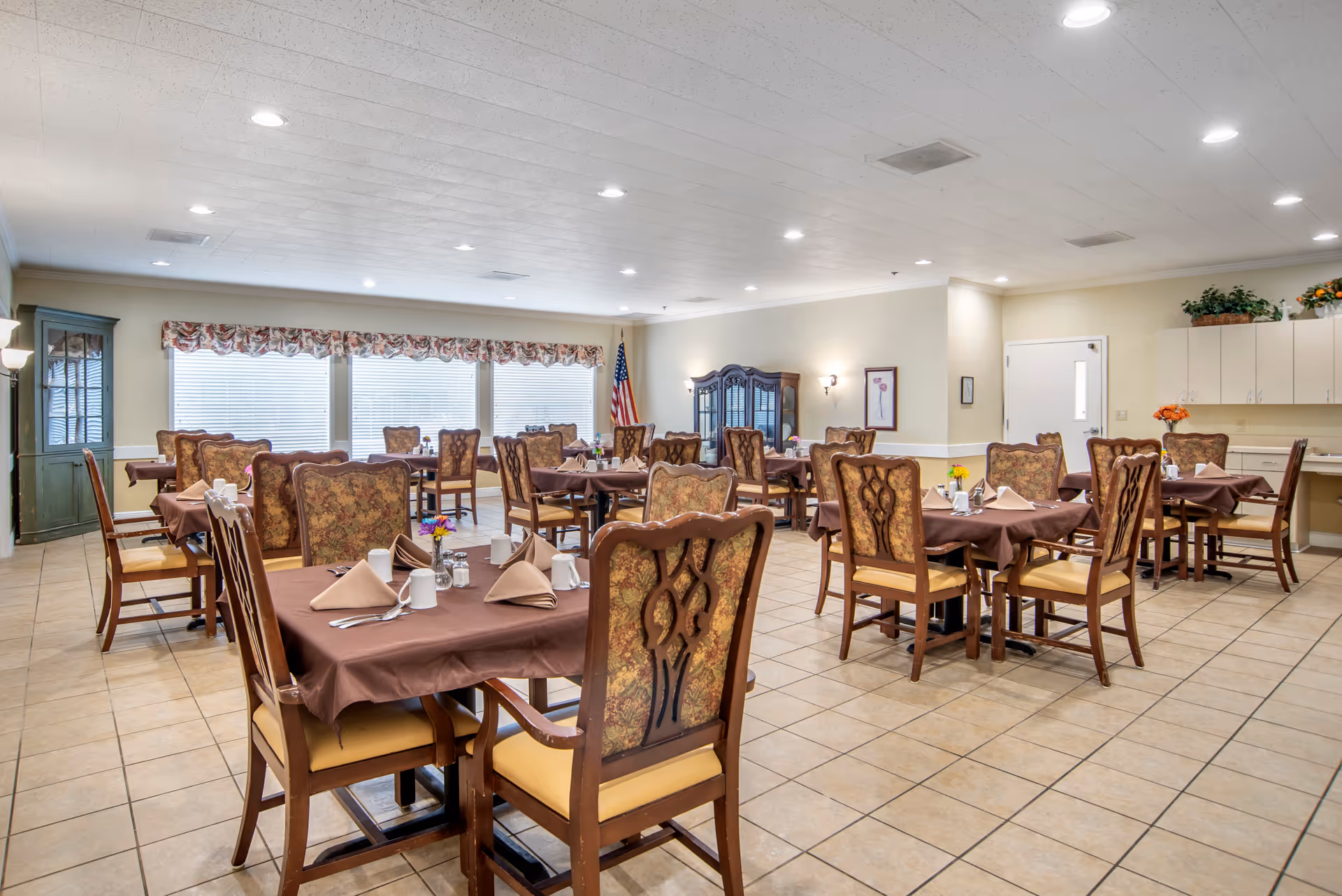 A spacious dining room with multiple tables covered in brown tablecloths, each set with beige napkins, white mugs, and silverware. The chairs have wooden frames with patterned upholstery. Large windows with floral valances let in natural light. The room has tiled floors, light-colored walls, and ceiling lights. There is an American flag in the corner and a cabinet with glass doors along the wall.