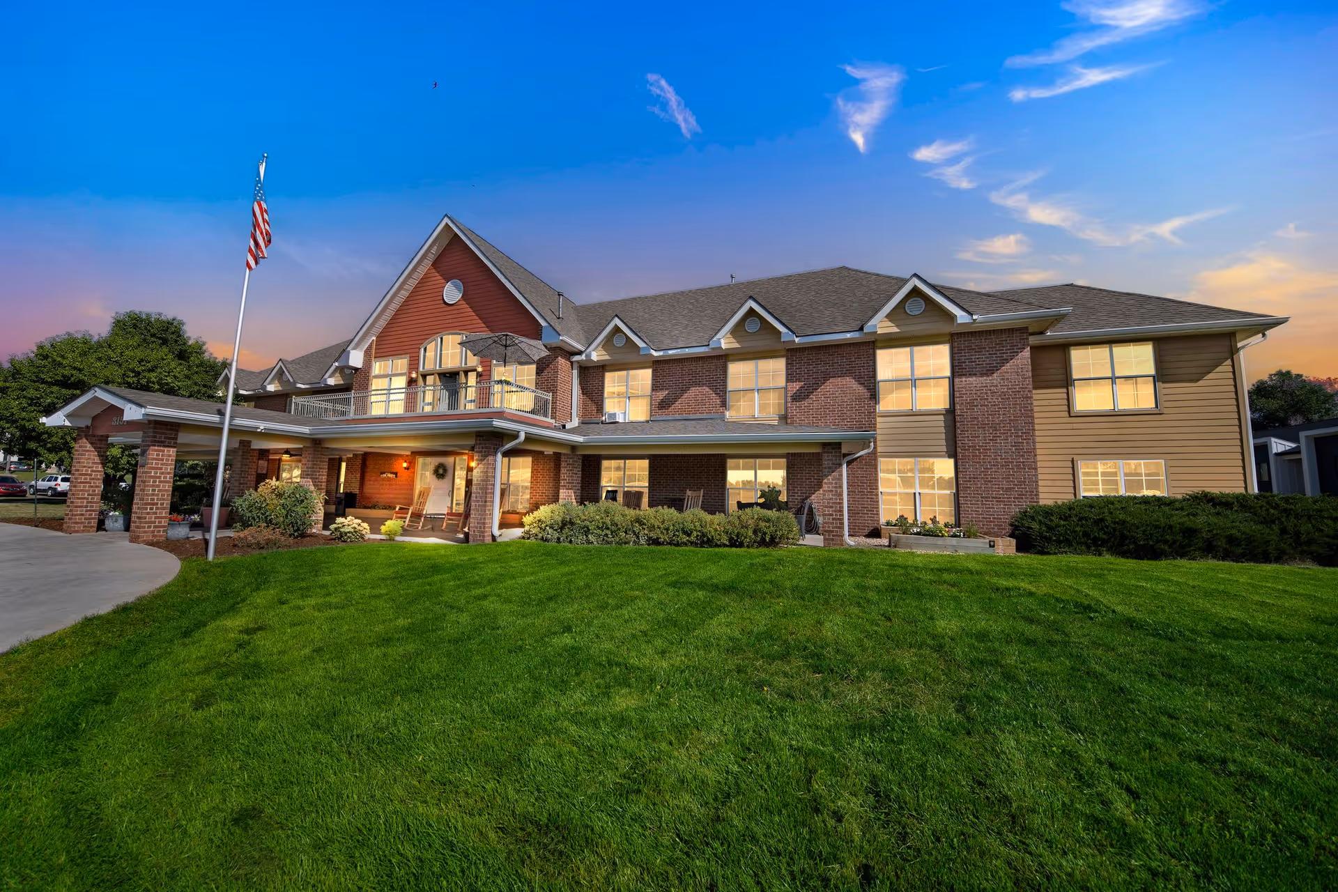 Exterior view of a two-story assisted living facility building with a well-maintained green lawn in front, an American flag on a flagpole, and a partly cloudy sky at sunset.