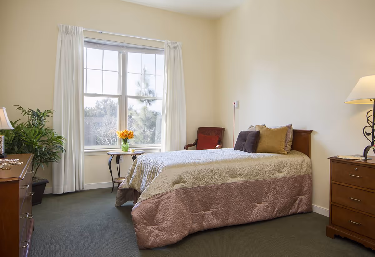 A cozy bedroom with a single bed covered in a beige and pink quilt, adorned with multiple pillows. There is a wooden dresser with a lamp and some items on top, a small round table with a vase of yellow flowers near a large window with white curtains, and a red cushioned chair in the corner. The room has light-colored walls and a green carpeted floor.