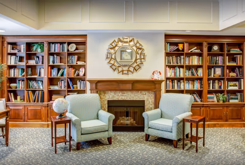 A cozy senior living facility common area featuring two light blue upholstered armchairs with wooden legs, each accompanied by a small wooden side table. Behind the chairs is a wooden fireplace with a decorative mirror above it. Flanking the fireplace are two large wooden bookshelves filled with books and decorative items. The floor is carpeted with a patterned design.