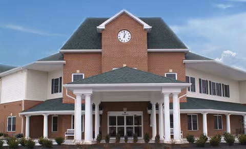Front exterior view of a senior living facility named The Birches at Harleysville, featuring a large brick building with a clock tower, white columns at the entrance, and a green roof under a partly cloudy sky.