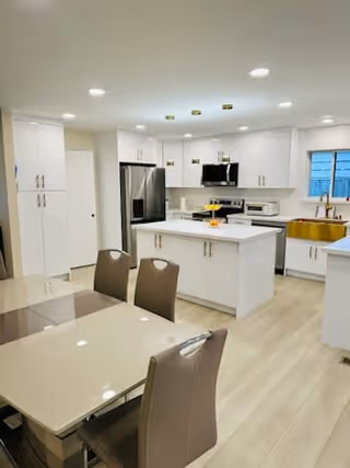 Open modern kitchen with white cabinets, a central island, stainless steel appliances, and a dining table in the foreground.