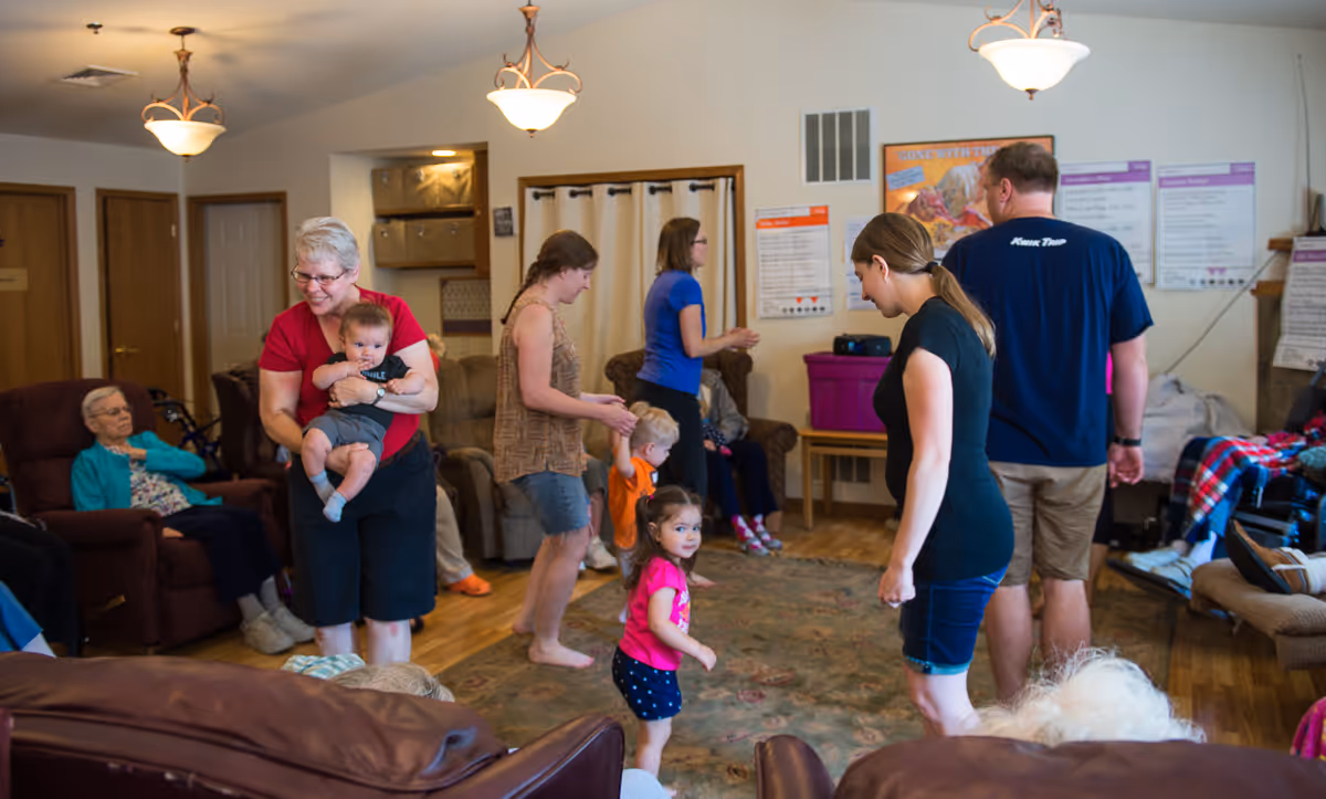 A lively common room in a senior living facility where elderly residents are seated on armchairs around the room while several adults and young children interact and move around the center of the room on a large rug. The room has warm lighting and a cozy atmosphere with wooden floors and light-colored walls.