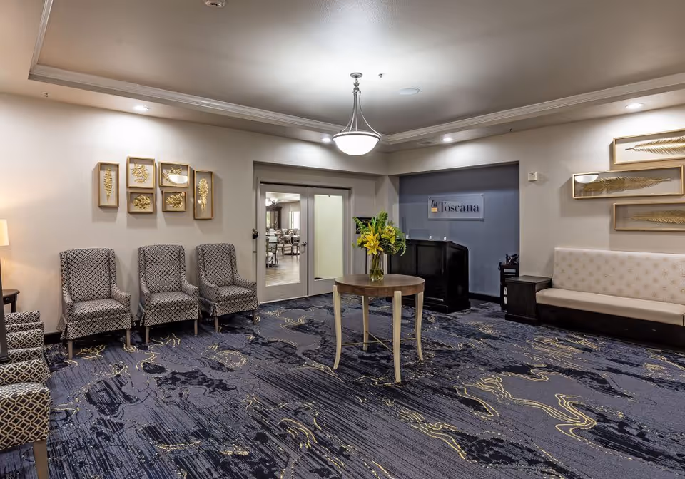 A spacious and well-lit common area with patterned carpet, several upholstered chairs arranged along the walls, a round wooden table with a vase of yellow flowers in the center, and decorative framed leaf artwork on the walls. Double glass doors lead to another room labeled 'la Toscana'.